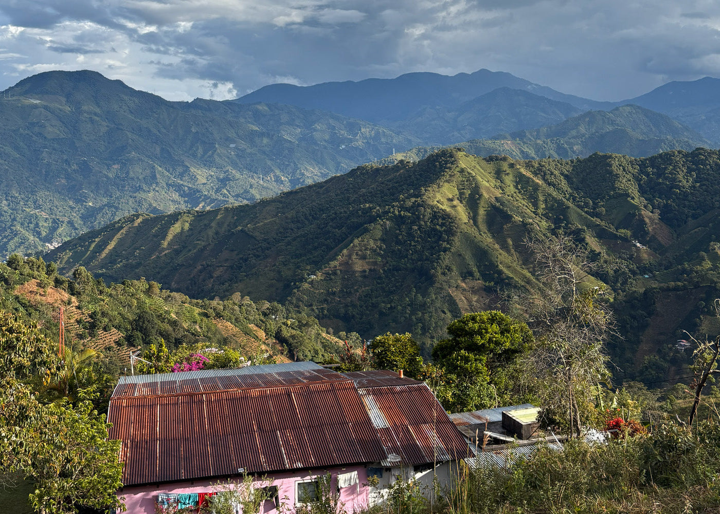 Hillside village with a rusty metal roof amidst green mountains under a cloudy sky.