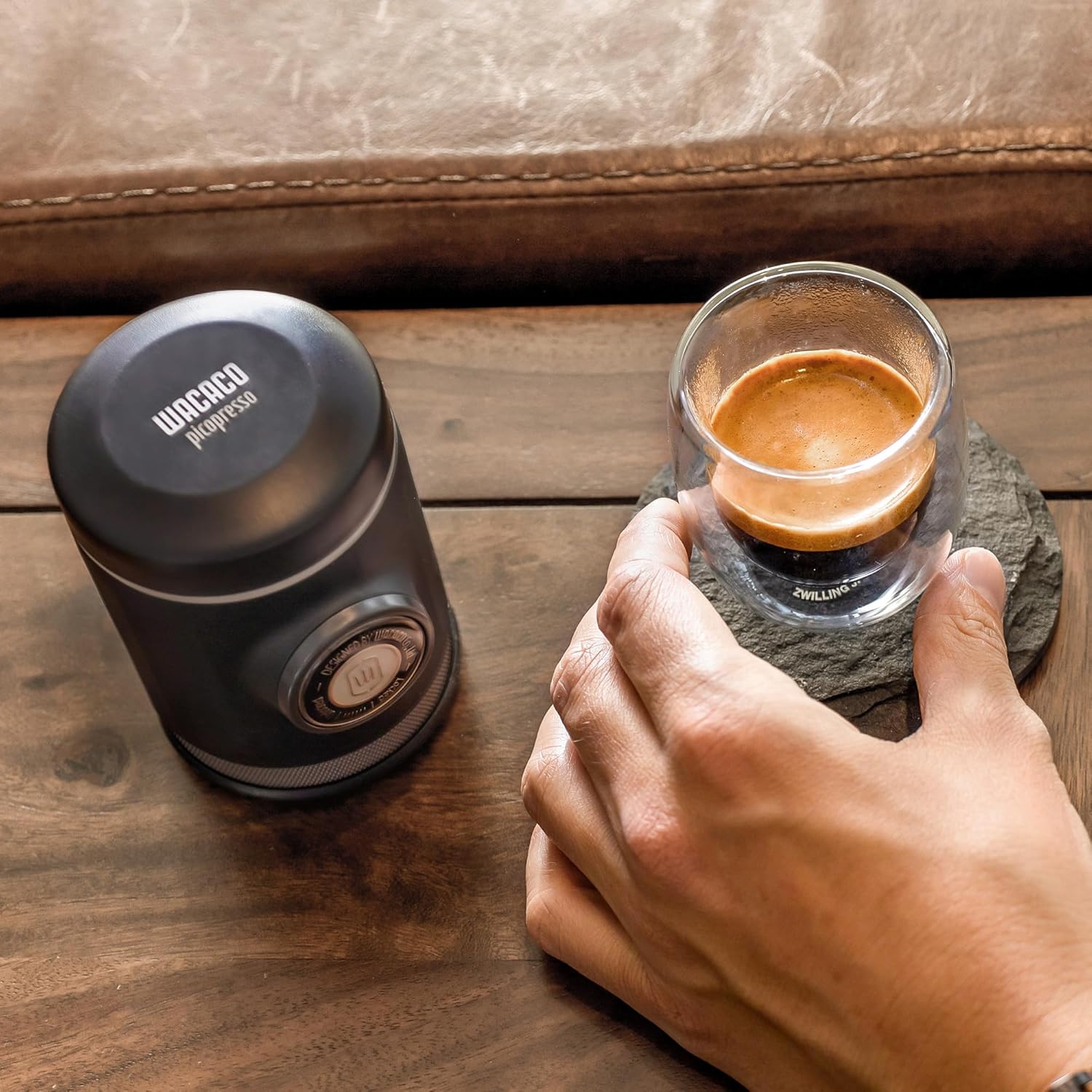Hand holding a glass of espresso next to a WACACO coffee maker on a wooden surface